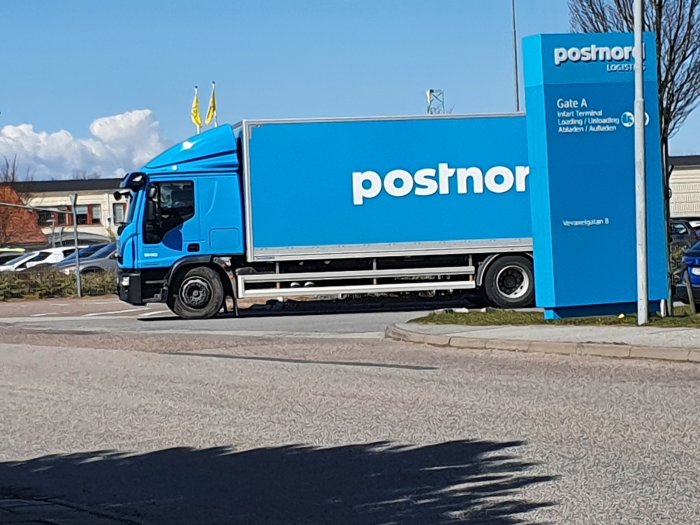 Blå lastbil med texten "poshnor" parkerad vid en terminal med skylten "Gate A".