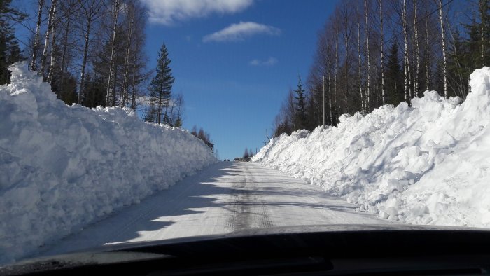 Väg omgiven av höga snövallar och skog sett från bilens förarsäte under en solig dag.