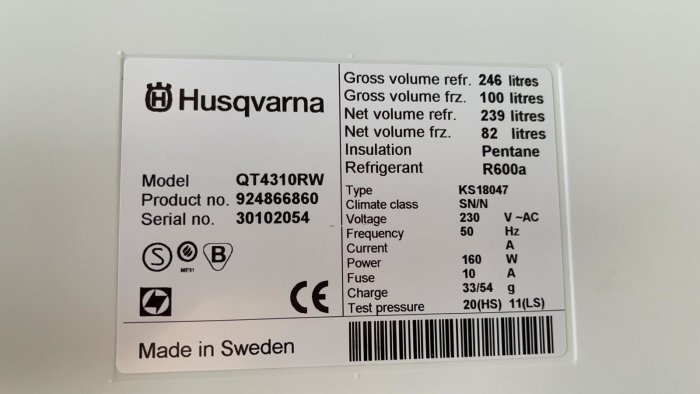 Typskylt för en Husqvarna-apparat med modellnummer, serienummer och specifikationer, fäst på en vit bakgrund.