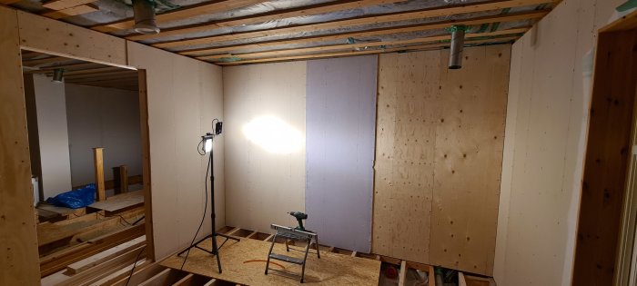 Under-konstruktion av badrum och bastu med oskyddade träreglar och plywood, byggbelysning och verktyg syns.