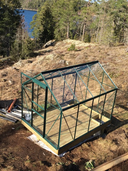 Påbyggnadsstadiet av ett grönt växthus med trägolv på en sommarstuga, omgiven av skog och med en sjö i bakgrunden.