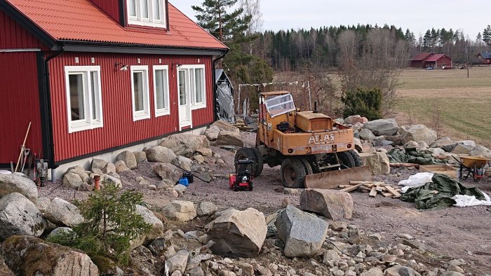 Byggplats vid röd stuga med stora stenar, en hjullastare och byggmaterial, planerad för altanbygge.