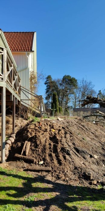 En slänt med uppgrävd jord vid ett hus, med trädgårdsredskap och träd i bakgrunden under en klarblå himmel.