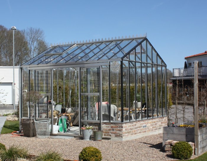 Växthus med glasstruktur på betongfundament med omgivande trädgård och bostadshus i bakgrunden.