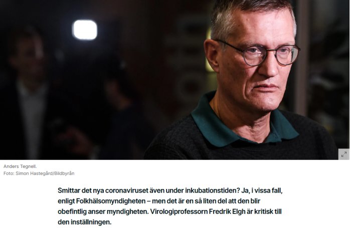 Man med glasögon ger en kritisk blick i ett suddigt rum, möjligt intervju eller presskonferens.
