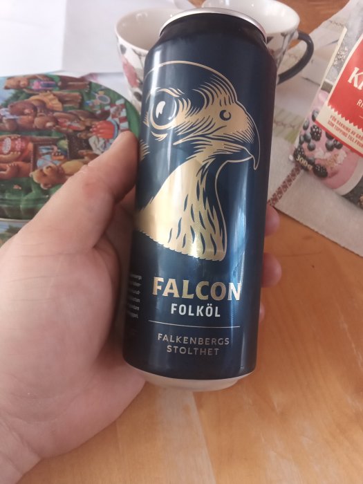 Hand håller en burk Falcon folköl med texten "Falkenbergs stolthet" på ett bord med tidningar och koppar i bakgrunden.