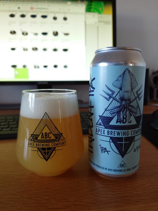 Ett glas öl bredvid en ölburk från Apex Brewing Company, synlig framför en datorskärm.