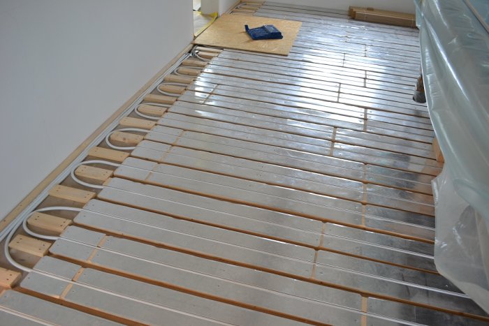 Golv med installerad golvvärme och armering, träreglar och metallstrålar synlig framför vägghörn och plastskydd.