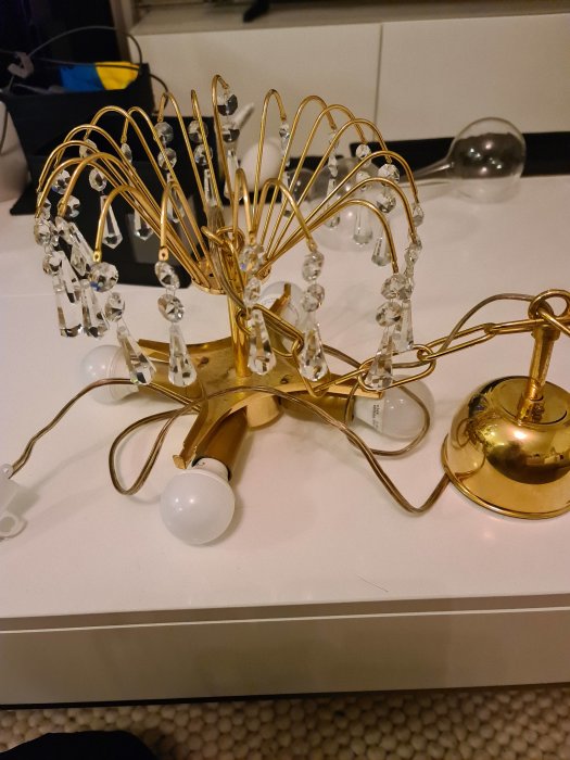 Guldfärgad armatur med genomskinliga kristaller och en vit glödlampa, utan märketikett, på en vit yta.