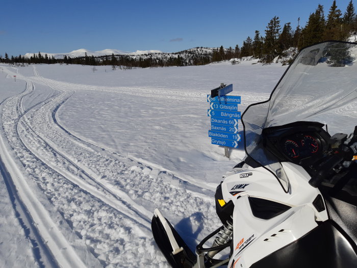 Snöskoter vid en vägvisare i ett vinterlandskap med skidspår och fjäll i bakgrunden.