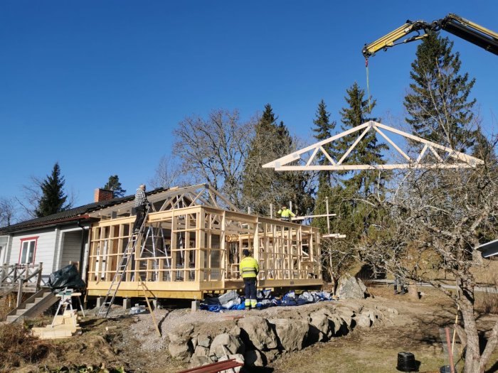 Installation av takstolar på ett husbygge med en kranbil, arbetare och ställningar i soligt väder.