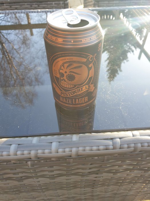 En ölburk märkt 'Pistonhead Haze Lager' med Galaxy-humle på ett reflekterande glasbord utomhus.