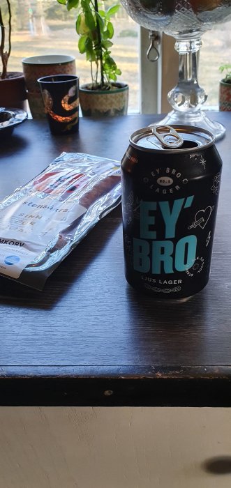 Ölburk med texten "EY BRO" på ett bord bredvid en förpackning rökt korv och bakomväxande krukväxter.