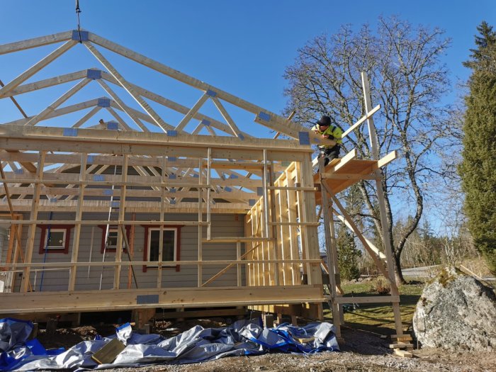 Arbetare monterar träkonstruktion på taket av ett hus under konstruktion, med klarblå himmel i bakgrunden.