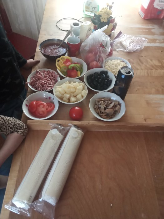 Ingredienser för hemgjord pizza på köksbord, med tomatsås, ost, skinka, grönsaker och deg.