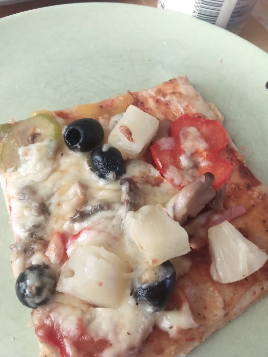 En bit pizza med oliver, ananas, svamp och paprika på en tallrik som symboliserar "fuskpizza".