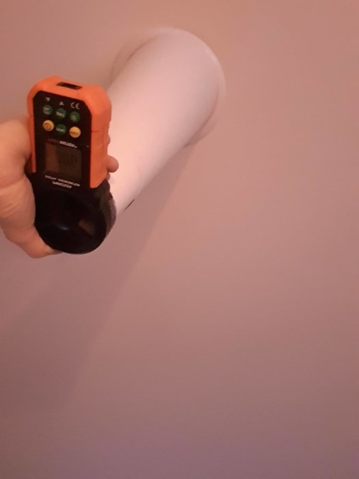 Hand håller en anemometer framför ett vitt ventilationsrör på en vind för att mäta luftflödet.