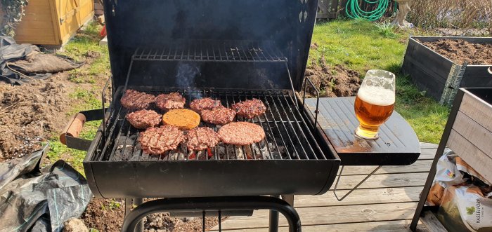 Hamburgare grillas på en kolgrill med ett glas öl vid sidan, utomhus på en solig dag.