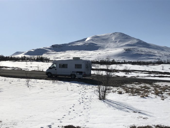 Husbil parkerad på snötäckt landskap med berg i bakgrunden under en klarblå himmel.