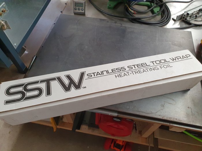 En låda märkt "STAINLESS STEEL TOOL WRAP - HEAT-TREATING FOIL" på ett arbetsbord i en verkstad.