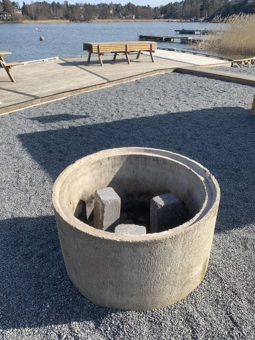 Beton grillring placerad på grus nära en sjö med bänkar och brygga i bakgrunden.