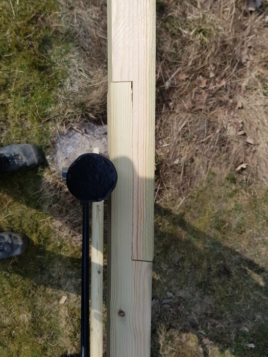 Två trästolpar med ett nyborrat 11 mm hål och skarv; det ena skarvat längre än det andra, mot gräsmark bakgrund.