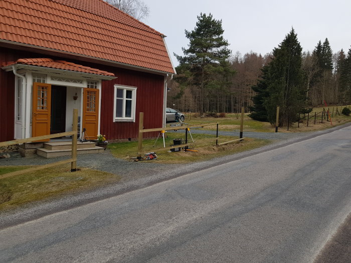Röd stuga med nya stolpar för en staketkonstruktion längs en lutande terräng vid en landsväg.