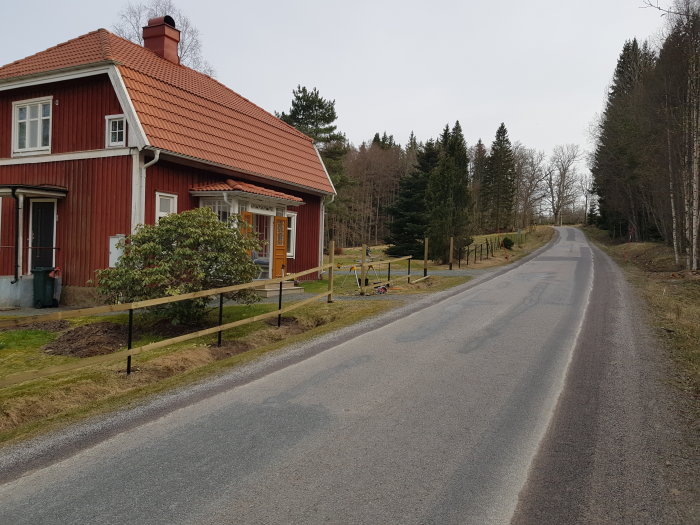 Röd stuga med nybyggt staket längs en ojämn väg i en lantlig miljö.