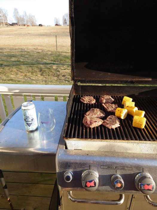 Grillning av hamburgare och majskolvar på en gasolgrill med en landskapsvy i bakgrunden.