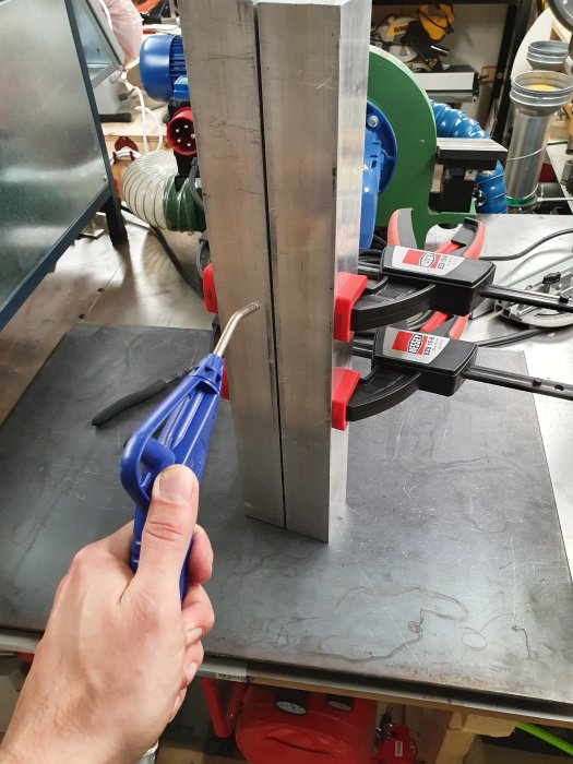 Hand håller en tång nära ett metallblad monterat i skruvstäd på en arbetsbänk.