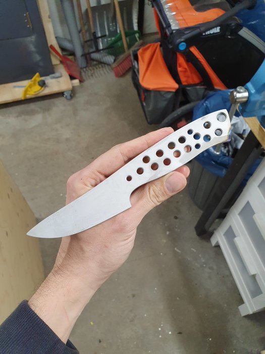 En hand håller upp en obehandlad kniv med hålmönster, mot en bakgrund av en verkstad.
