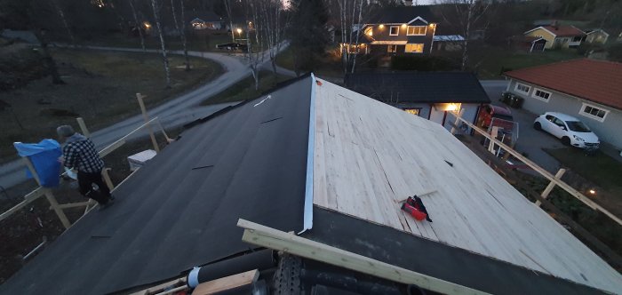 Tak med råspont och delvis monterad takpapp, personer arbetar, i ett bostadsområde vid skymning.