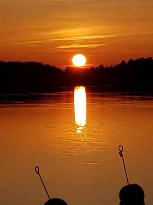 Solnedgång över en stilla sjö med reflektion av solen i vattnet, konturer av två fiskespön i förgrunden.