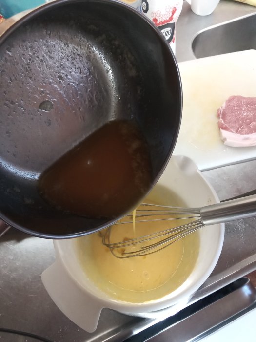 Hällning av smält smör i skål med äggula för hemmagjord bearnaisesås, visp och rå fläskstek i bakgrunden.