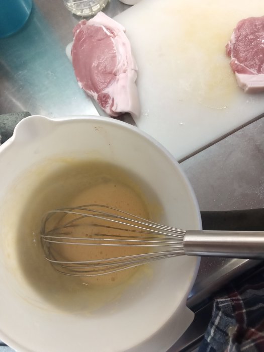 Beredning av hemmagjord bearnaisesås med visp i skål, rå fläskstek i bakgrunden.