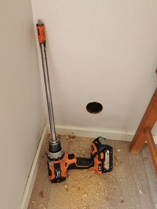 En borr med långt borrskär i hörn av rum vid hål i golv, omgiven av sågspån.