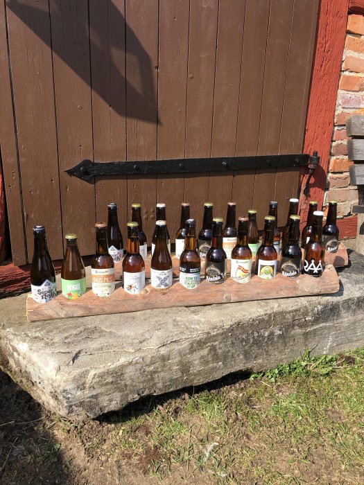 Samling av olika ölflaskor från skånska bryggerier uppställda på en sten framför en röd trävägg.