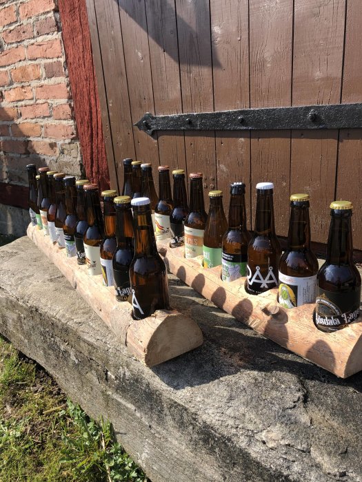 Samling av olika ölflaskor från skånska bryggerier uppställda på en stenmur i solljus.