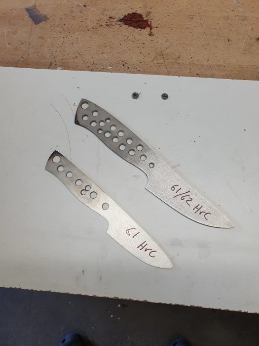 Två oslipade knivblad i metall med utsågade hål, märkta med hårdheten 61 HRC respektive 61½ HRC, på en arbetsbänk.