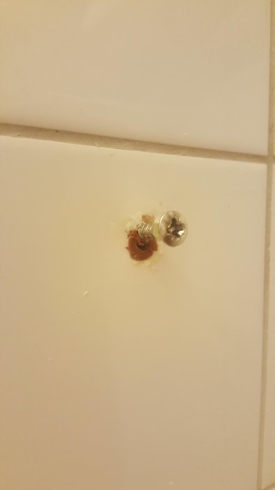 Skruv i badrumskakel med missad plugg och synligt hål som tidigare dolts av duschstångshållare.