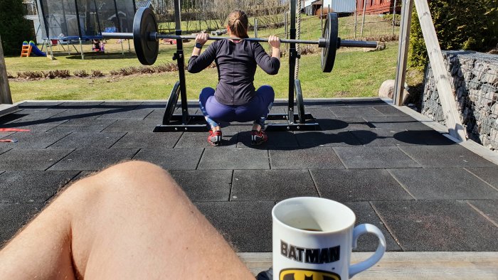 Person utför knäböj med skivstång utomhus, observerad över en kaffekopp med Batman-logga.