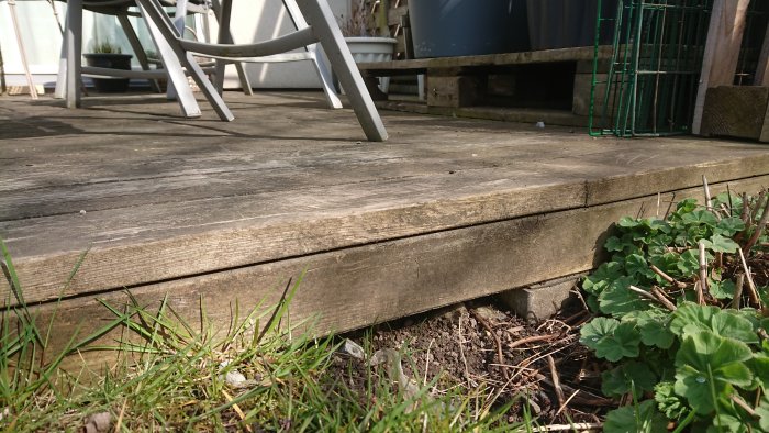 Trädäck nära marken med synliga gräs och blad, del av kompostgaller och trädgårdsmöbler i bakgrunden.