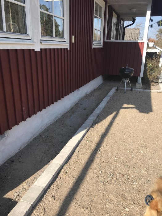 Nybyggd stenkant längs med husgrund och grusyta, med en klotgrill och hund i hörnet.