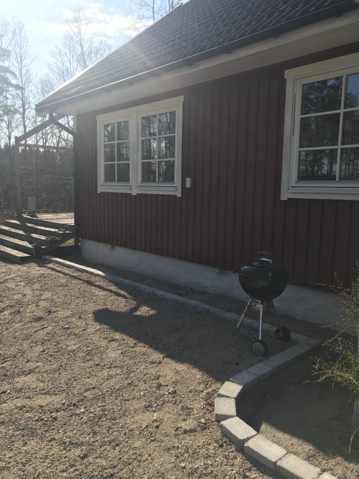 Röd trähusfasad med vita fönster, grusad mark framför huset och en kolgrill på betongplattor.