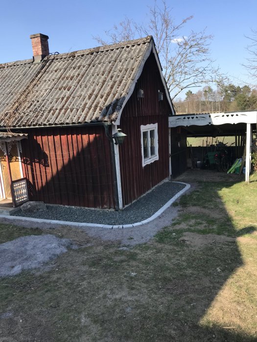 Grusgång med kantsten längs en röd stuga och dess uthus, med synliga ytor där vegetation tagits bort.