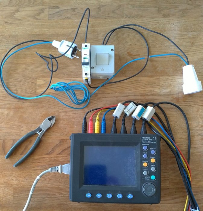 Elektrisk testuppställning med mätinstrument, kablar, tänger och kopplingar på ett trägolv.