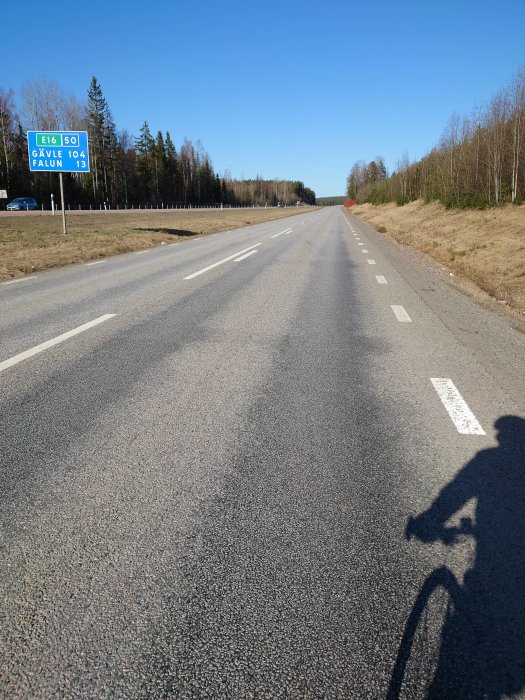 En tom landsväg med skylt för E16, och skugga av en cyklist till höger.