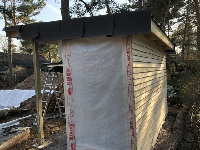 Hörnet av en liten byggnad under konstruktion med spikläkt och fjällpanel, omgiven av trädgård och byggmaterial.