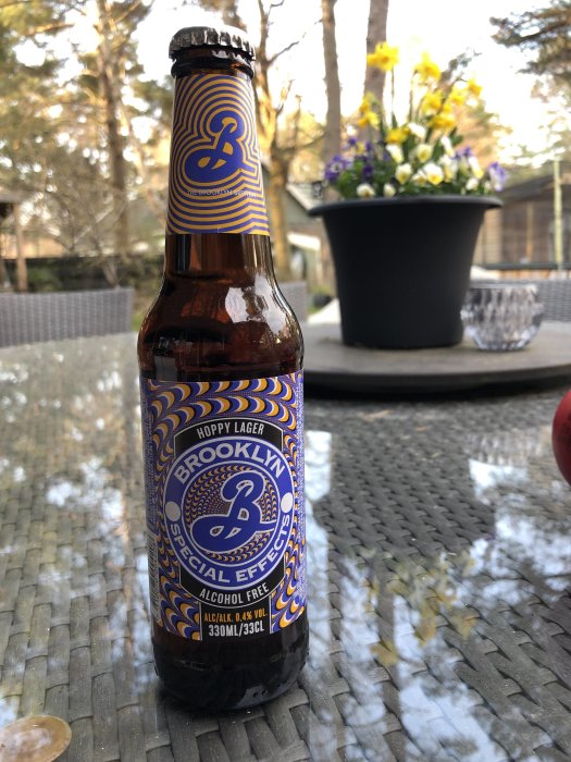 Flaska Brooklyn Special Effects alkoholfri öl på bord utomhus, suddig blomkruka i bakgrunden.