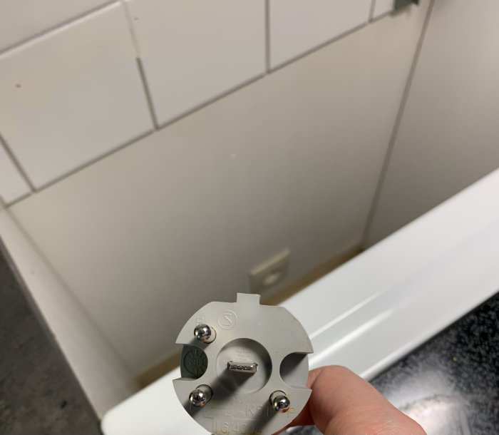 Hand håller en grå spis-kontakt framför en vit kaklad vägg och köksbänk, frågar om el-uttagskompatibilitet.